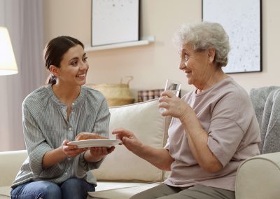 A caregiver with an elderly woman helping with her meal on the sofa