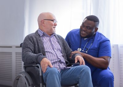 an elderly man in a wheelchair with an African American caregiver both smiling